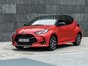 Een levendige rode Yaris-hybride geparkeerd op een betonnen oppervlak tegen een grijze stenen muur straalt moderne elegantie uit.