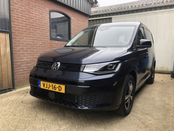 Buiten staat een zwart Volkswagen Caddy-busje geparkeerd, voor een bakstenen gebouw en garage, met een gele kentekenplaat met de tekst "VJJ-16-D".