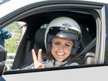 Persoon die een racehelm en -pak draagt, in een auto zit, glimlacht en met de hand een vredesteken gebaar maakt. Het is alsof Sabine Schmitz zelf achter het stuur zit.