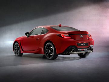 Een strakke, rode Toyota GR 86 sportwagen met achterspoiler en dubbele uitlaten, van achteren gezien, geparkeerd in een minimalistische ruimte met betonnen muren.