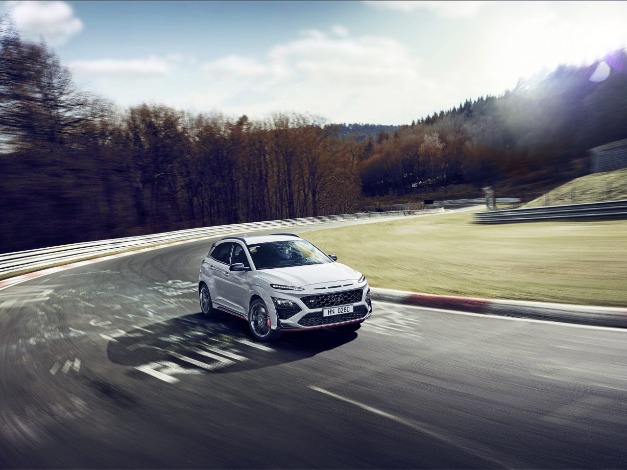 Een witte Hyundai Kona N SUV rijdt op een racecircuit met herfstbomen op de achtergrond onder een gedeeltelijk bewolkte hemel.