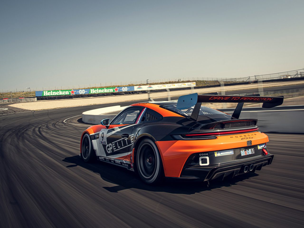 Een zwart-oranje 911 GT3 Cup-auto met het nummer 91 snelt over het circuit. Reclamebanners en een vangrail omlijsten de achtergrond.