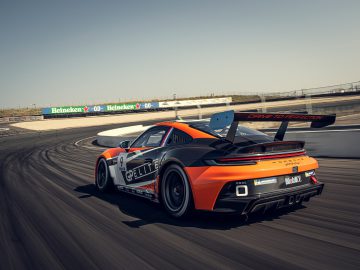 Een zwart-oranje 911 GT3 Cup-auto met het nummer 91 snelt over het circuit. Reclamebanners en een vangrail omlijsten de achtergrond.
