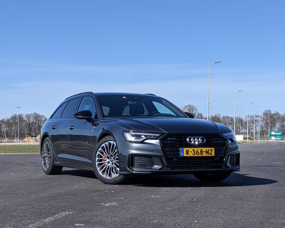 Een donkergrijze Audi A6 Avant stationwagen staat geparkeerd op een asfaltoppervlak onder een helderblauwe hemel. De auto heeft een geel Nederlands kenteken met nummer K-368-NZ. Op de achtergrond zijn bomen zichtbaar.