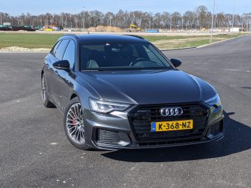 Een grijze Audi A6 Avant met Nederlands kenteken K-368-NZ staat geparkeerd op een asfaltoppervlak, met op de achtergrond bomen en een bouwplaats.
