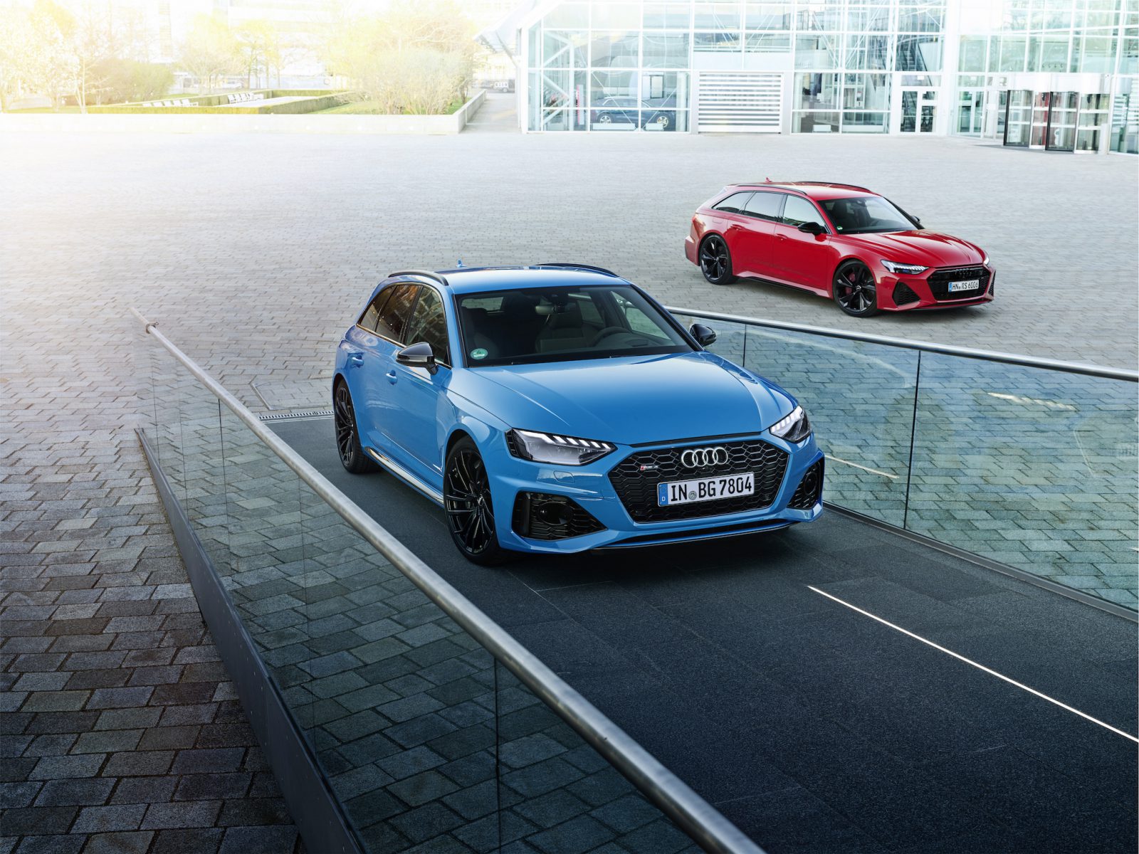 Twee Audi Sportwagens, een blauwe en een rode, staan geparkeerd op een moderne buitenruimte met een glazen balustrade en een groot gebouw op de achtergrond.