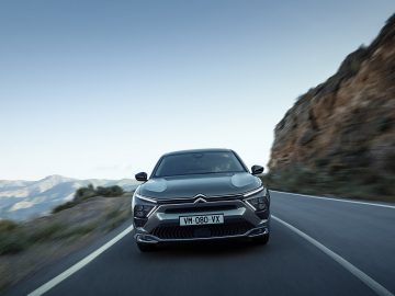 Een strakke zwarte Citroën C5 X met kenteken "VM-080-YK" glijdt over een kronkelende bergweg, kliffen aan de rechterkant en majestueuze bergen die als adembenemende achtergrond dienen.