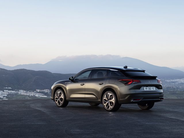 Een strakke, grijze Citroën C5 X staat geparkeerd op een hoge, verharde ondergrond met uitzicht op een uitgestrekt landschap met bergen en een verre stad. De scène vindt plaats onder een heldere hemel in de schemering.