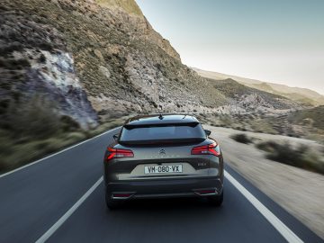 Een grijze Citroën C5 X rijdt op een bochtige bergweg met een landschap van rotsachtige heuvels en een heldere hemel op de achtergrond. Op het kenteken van de auto staat "VM-080-XX".