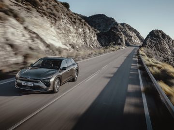 Een zwarte Citroën C5 X SUV rijdt op een kronkelige bergweg onder een heldere hemel, omgeven door rotsachtige heuvels en schaarse vegetatie.