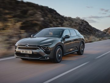 Een donkergrijze Citroën C5 X SUV rijdt op een gladde weg met een wazig woestijnlandschap en rotsachtige heuvels op de achtergrond onder een gedeeltelijk bewolkte hemel.