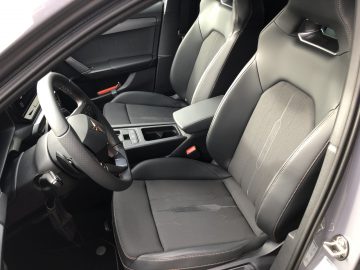 Interieur van een moderne Cupra Leon e-Hybrid met strakke zwartleren voorstoelen, middenconsole met opbergruimte en versnellingspook, en een stijlvol stuurwiel. De deur staat open en biedt vrij zicht op de bestuurderszijde.