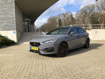 Op een verhard terrein voor een gebouw met trappen en grote ramen staat een grijze Cupra Leon e-Hybrid met geel kenteken geparkeerd. Bomen zijn zichtbaar op de achtergrond onder een gedeeltelijk bewolkte hemel.