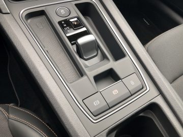 Close-up van de middenconsole van de Cupra Leon e-Hybrid, met de versnellingspook, de parkeerremknop, de automatische start/stop-knop en andere bedieningselementen. Het interieur is zwart met witte stiksels.