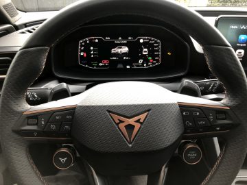 Close-up van het stuur van een Cupra Leon e-Hybrid met een digitaal dashboarddisplay met een voertuigafbeelding, verschillende bedieningselementen en een start/stop-knop aan de rechterkant.