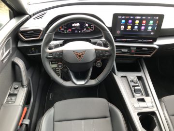 Binnenaanzicht van de cockpit van de Cupra Leon e-Hybrid met een stuurwiel, digitaal dashboard, infotainmentsysteem met touchscreen en moderne bedieningselementen op de middenconsole en deur.