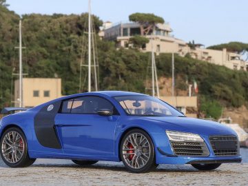 Op een stenen ondergrond staat een blauwe sportwagen geparkeerd met op de achtergrond een jachthaven en huizen op een heuvel, wat lijkt op een ingewikkeld schaalmodel tafereel.