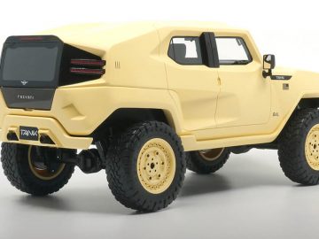 Een beige, stoere terreinwagen met grote banden en een strak, hoekig ontwerp wordt tentoongesteld in een goed verlichte studiosetting, vergezeld van een gedetailleerd schaalmodel.