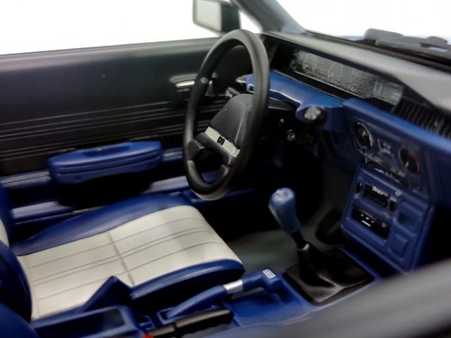 Interieur van een auto met een stuur, handmatige versnellingspook en blauwgestreepte stoelen met een donker dashboard en deurpanelen. Het uitzicht is vanaf de bestuurderszijde, alsof je de ingewikkelde details van een levensgroot schaalmodel bekijkt.