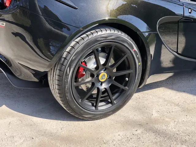 Close-up van het achterwiel van een zwarte Lotus Elise met een zichtbare rode remklauw, geparkeerd op een betonnen ondergrond.