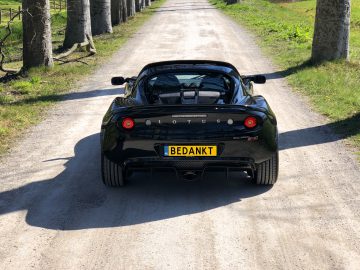 Een zwarte Lotus Elise met een "BEDANKT" kentekenplaat staat op een zonnige dag geparkeerd op een grindpad geflankeerd door bomen.