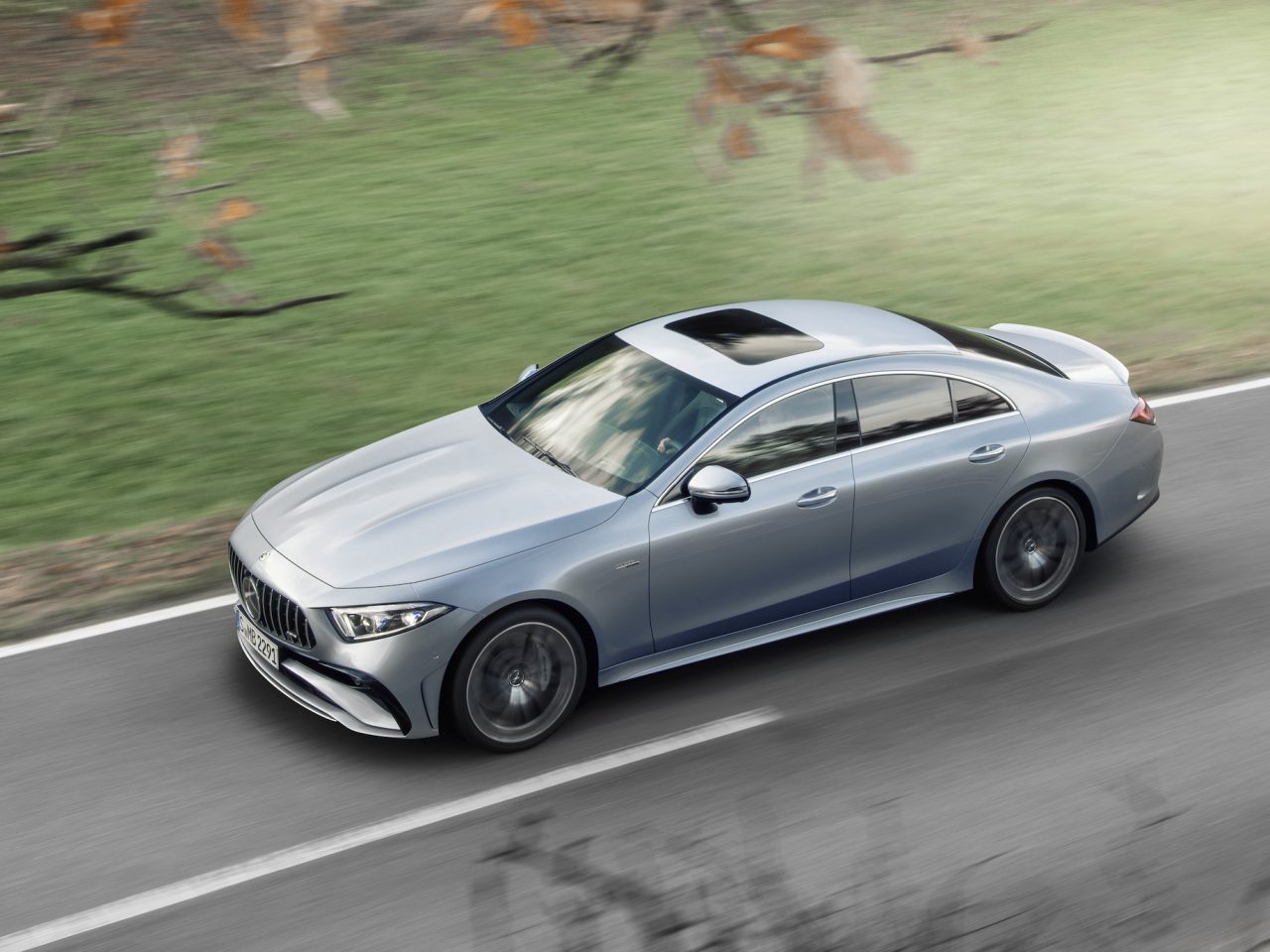 Een zilveren Mercedes-Benz CLS met schuifdak rijdt over een verharde weg, omringd door een grasveld en enkele bomen op de achtergrond.