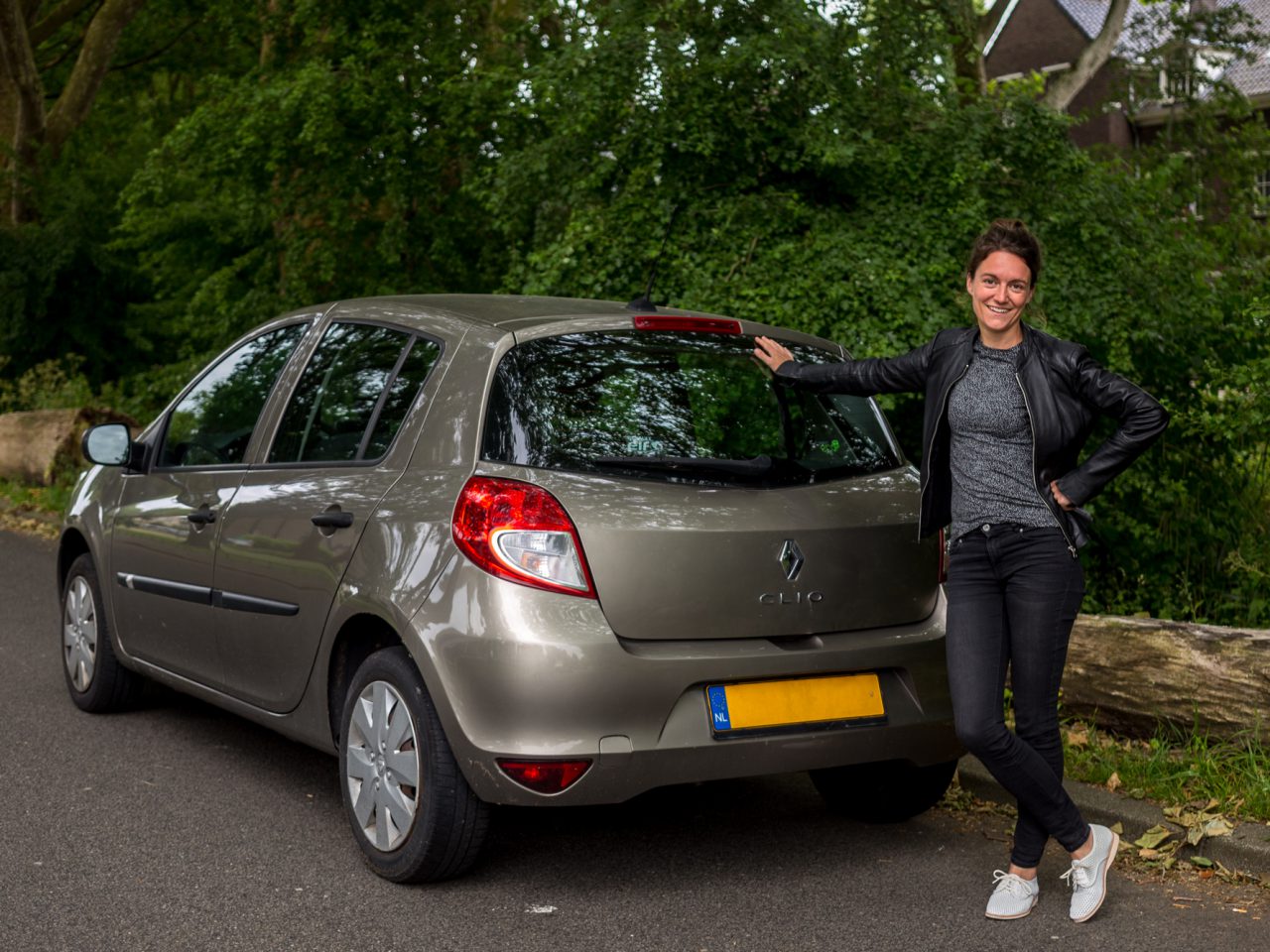 Een persoon staat naast een grijze Renault Clio, verworven via private lease, geparkeerd aan een weg met bomen en een gebouw op de achtergrond.
