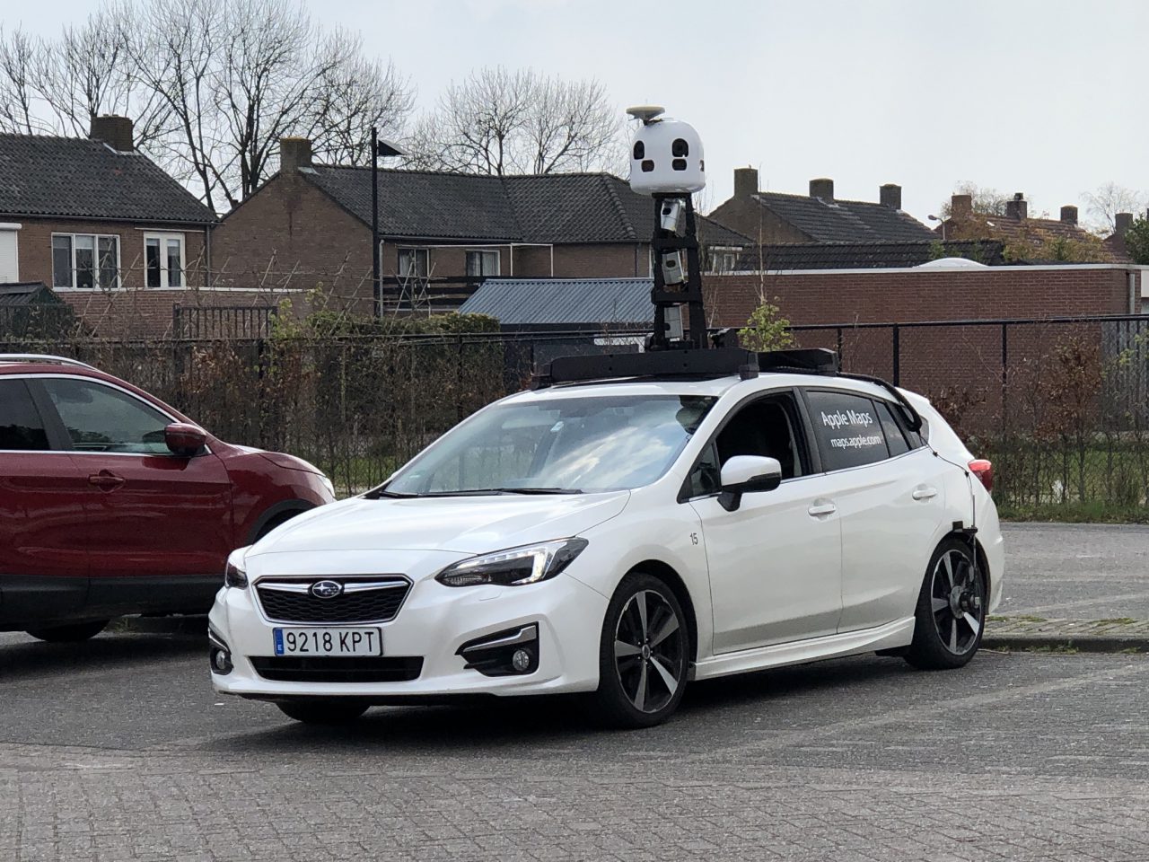 Een witte Subaru-auto uitgerust met een hoog camerasysteem op het dak staat geparkeerd in de buurt van rode en bruine gebouwen, met een bord aan de zijkant met de tekst 'Apple Maps.