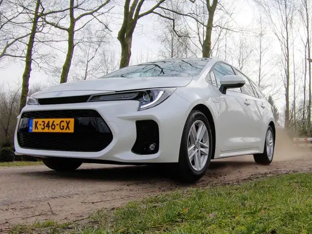 Een witte Suzuki Swace sedan met Nederlands kenteken K-346-GX rijdt op een onverharde weg omringd door bladerloze bomen.