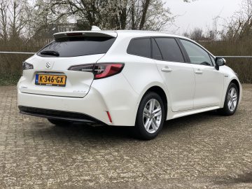 Op een verharde ondergrond met bomen op de achtergrond staat een strakke, witte Suzuki Swace hybride auto met Nederlands kenteken K-346-GX geparkeerd.