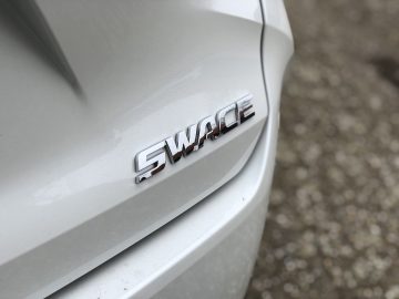 Close-up van het "Suzuki Swace" -embleem op een wit voertuig, gefocust op het achterste gedeelte van de auto, terwijl de achtergrond een gestructureerd grondoppervlak laat zien.