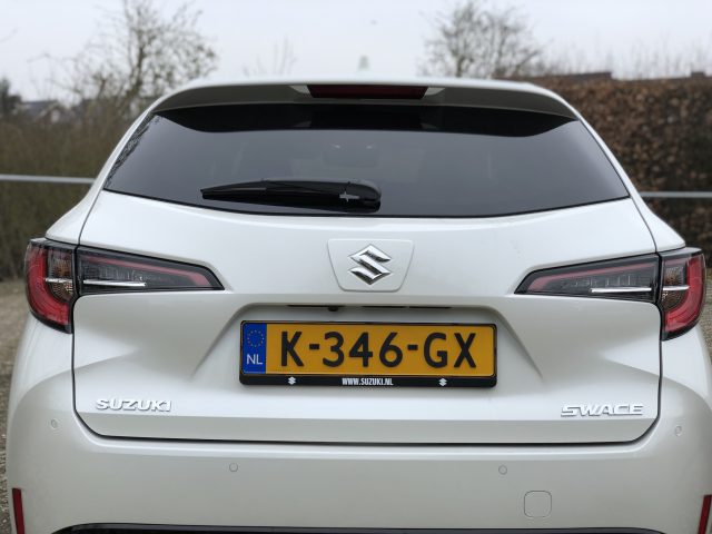 Achteraanzicht van een witte Suzuki Swace met een Nederlands kenteken met de tekst "K-346-GX." De auto staat buiten geparkeerd met een hek en bomen op de achtergrond.