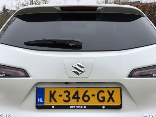 Achteraanzicht van een witte Suzuki Swace met Nederlands kenteken, K-346-GX, geparkeerd. De auto is voorzien van een ruitenwisser achter en het Suzuki-embleem.