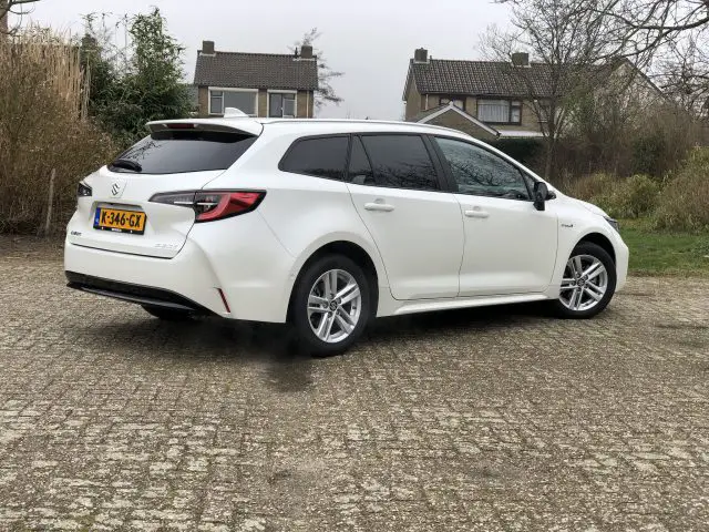 Op een verhard terrein staat een witte Suzuki Swace met Nederlands kenteken K-346-GX geparkeerd, met op de achtergrond woonhuizen en groen.