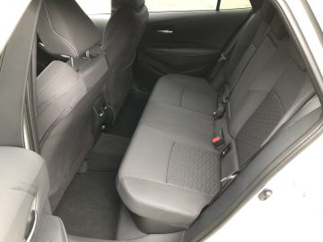 Het interieur van de Suzuki Swace onthult het achterbankgedeelte met grijze stoffen bekleding en stoelpatronen. De autodeur staat een stukje open, waardoor de ruime beenruimte en de netjes gerangschikte veiligheidsgordelgespen goed tot hun recht komen.