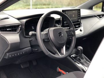 Interieur van een Suzuki Swace met een zwart dashboard met het Suzuki-logo op het stuur, een touchscreen in de middenconsole en verschillende bedieningsknoppen en versnellingspook.