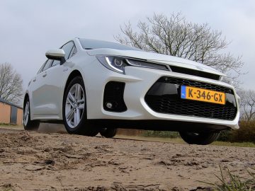 Een witte Suzuki Swace met een geel kenteken "K-346-GX" staat geparkeerd op een onverharde weg. Het is omgeven door bladerloze bomen en gebouwen onder een bewolkte hemel.