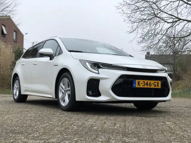 Op een verhard terrein nabij huizen en kale bomen staat een witte Suzuki Swace hatchback met Nederlands kenteken K-346-GX geparkeerd.