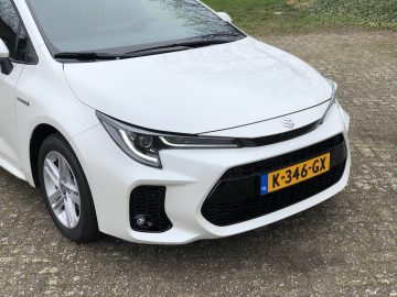 Een witte Suzuki Swace hybride auto geparkeerd op een gestructureerd oppervlak, met een Nederlands kenteken met het nummer K-346-GX.