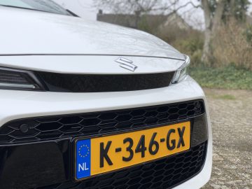 Close-up van de voorkant van een witte Suzuki Swace met een geel Nederlands kenteken met de tekst "K-346-GX" in een buitenomgeving met bomen en struiken op de achtergrond.