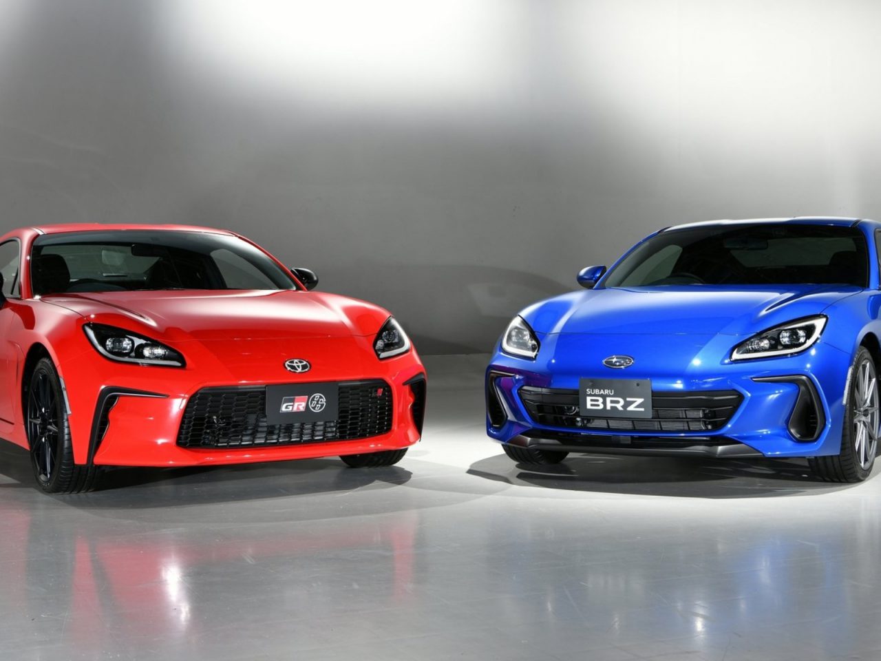 Een rode Toyota GR 86 en een blauwe Subaru BRZ-sportwagen staan binnen naast elkaar geparkeerd op een reflecterende vloer.