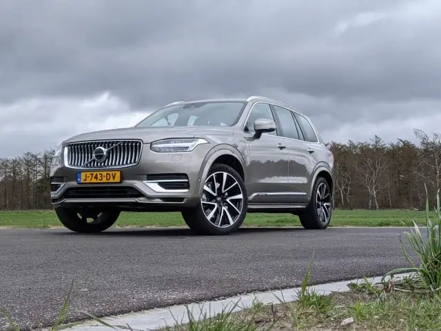 Een grijze Volvo XC90 met Nederlands kenteken J-743-DV staat geparkeerd op een asfaltweg omgeven door groen onder een bewolkte hemel.