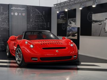 Een rode Abarth 1000 SP wordt tentoongesteld in een showroom met een decor met een erfgoedthema, inclusief afbeeldingen van oldtimers en blauwdrukken aan de muren. De verlichting benadrukt het strakke design van de auto.