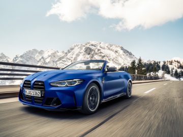 Een blauwe BMW M4 Competition Cabrio rijdt over een bergweg met besneeuwde bergtoppen op de achtergrond.