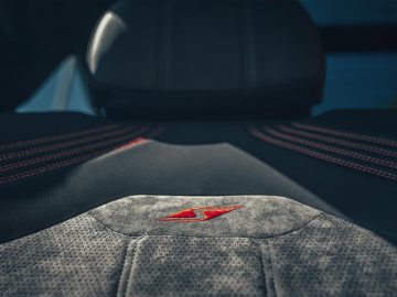 Close-up van een autostoeltje met zwarte en grijze stof, met rode stiksels en een geborduurd rood en wit "S"-logo in het midden, dat doet denken aan de luxueuze Bentley Bentayga S.