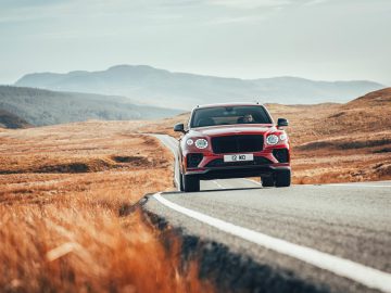 Een rode Bentley Bentayga S rijdt op een bochtige weg door een bergachtig landschap met grasvelden.