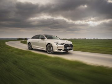 Een zilverkleurige DS 9 sedan glijdt over een bochtige weg door een graslandschap onder een bewolkte hemel.