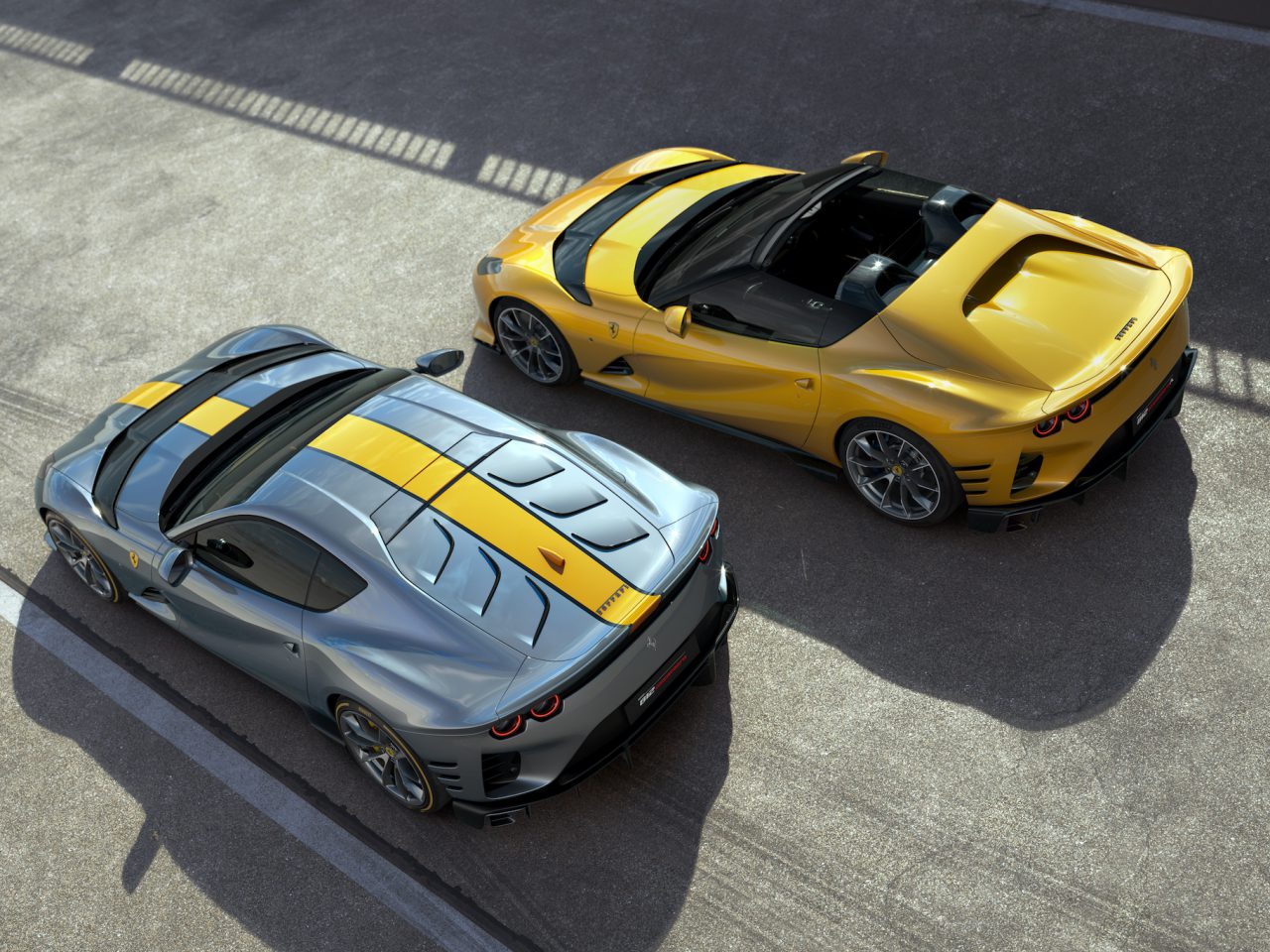 Twee hoogwaardige sportwagens, waaronder een Ferrari 812 Competizione in zilver met gele accenten en een andere in geel met zwarte accenten, worden vanuit een bovenaanzicht getoond. Ze staan geparkeerd op een betonnen ondergrond, met de schaduw van een hek ertussen.
