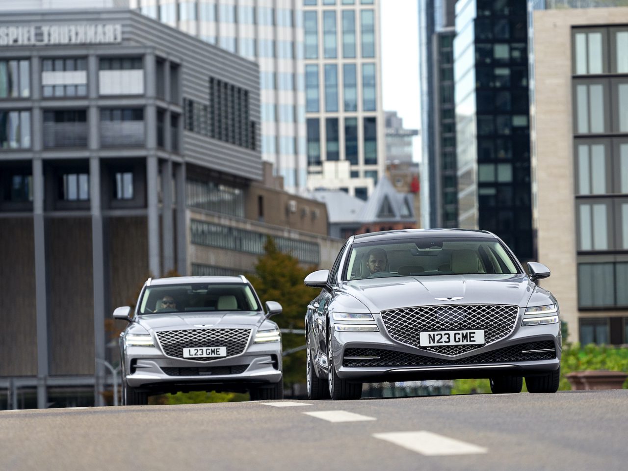 Twee zilveren Genesis-auto's met Britse kentekenplaten, "N23 GME" en "L23 GME", rijdend op een stadsweg met moderne gebouwen op de achtergrond.