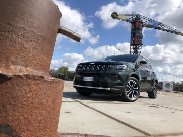 Een groene Jeep Compass SUV staat geparkeerd op een betonnen ondergrond nabij een verroeste metalen paal met een grote industriële kraan op de achtergrond onder een gedeeltelijk bewolkte hemel.